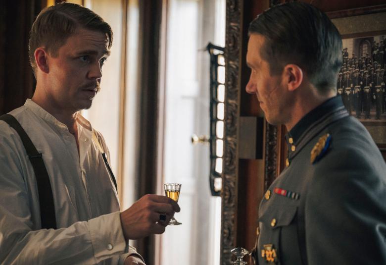 Kuvassa on vasemmalla Aku Sipola valkoisessa paidassa kohottamassa lasia sotilaspuvussa olevalla Jussi Vataselle, joka on osittain selin. Taustalla on iso ikkuna ja kuva sävy on tummahko. 