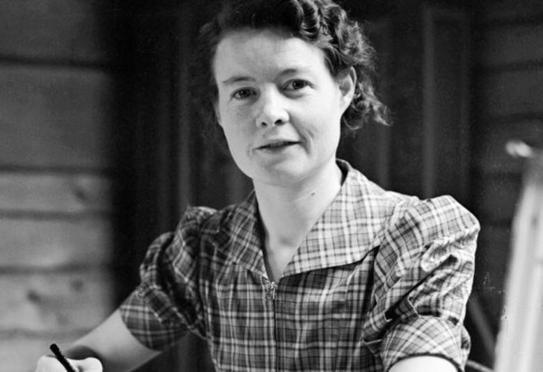 Kuvassa on Sally Salminen istumassa ja kirjoittamassa kynä kädessä. Hänellä on ruudullinen mekko päällään. Taustalla on hirsiseinää. Kuva on musta-valkoinen.