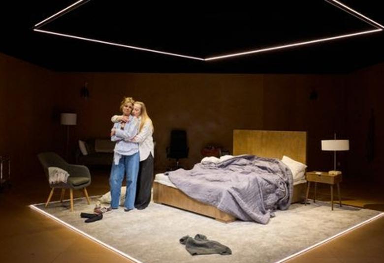 Kuvassa on valaistulla näyttämöllä, jossa on takana oleva pitkähiuksinen, valko-paitainen Milla Kangas halaa Anni-Maija Koskista, jolla on siniset farkut ja vaalean sininen paitapusero. Heidän takanaan on pari vuode, jossa on vaalean harmaa päiväpeitto sekaisin. 