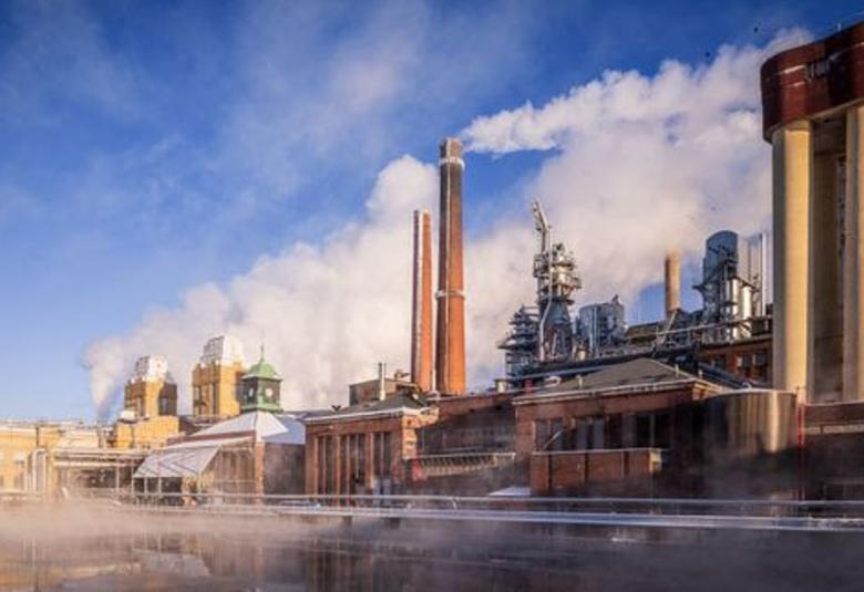 Kuvassa on edessä vettä ja takana punatiilisiä tehdasrakennuksia, joissa on torneja ja oikealla korkeat savupiiput, joista tulee savua. Tautalla on sinistä taivasta.