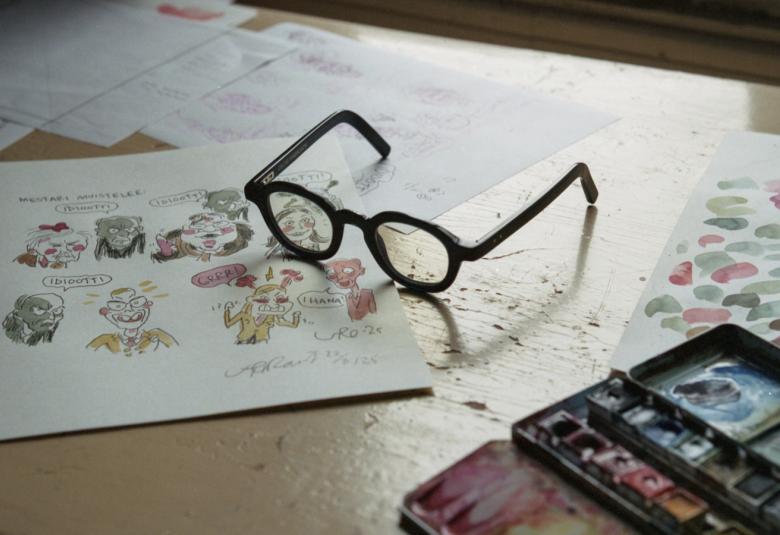 Kuvassa on pilapiirroskuvia pöydällä valkoisella paperilla ja niiden päällä silmälasit. Oikeassa reunassa on värejä.