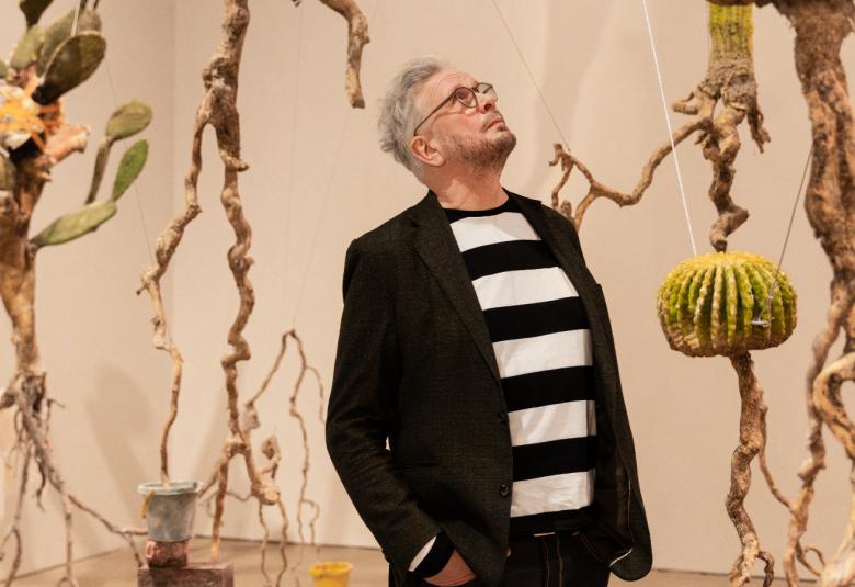 Kuvassa on keskellä Roland Persson seisomassa puolivartalokuvassa. Hänellä on musta takki ja musta-valkoinen paksuraidallinen pusero päällään. Hän katsoo ylöspäin. Taustalla on roikkuvia juurista tehtyjä taideteoksia ja seinät ovat vaalean ruskeat.