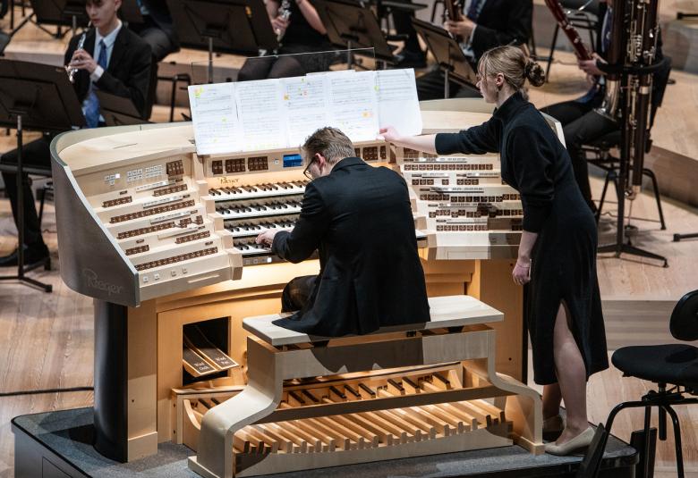 Kuvassa on keskellä selin soittamassa urkuja Aleksanteri Wallius ja urut ovat leveästi orkesterin edessä. Orkesteria näkyy hieman. Urkurin vieressä seisoo nainen, joka kääntää nuottia urkurille.