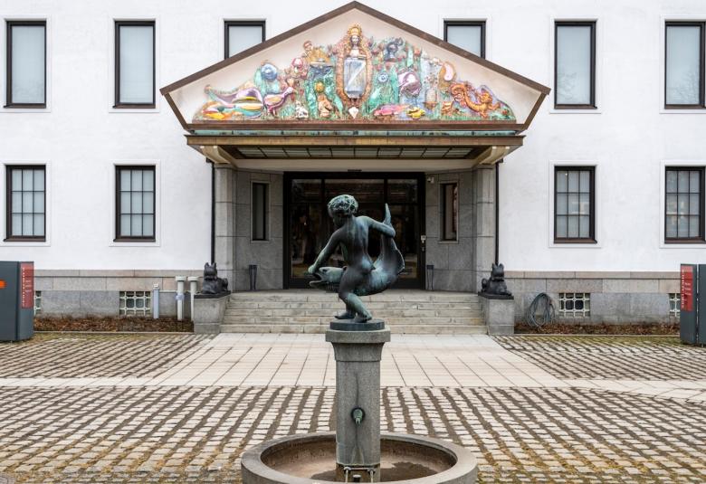 Kuvassa on valkoinen rakennus jossa on kapeita ikkunoita kahdessa kerroksessa. Siinä on portaikko ja isot ovet ja niiden päällä iso kolmio. Siinä on reliefimäisesti palstellivärisiä hahmoja ja esineitä.