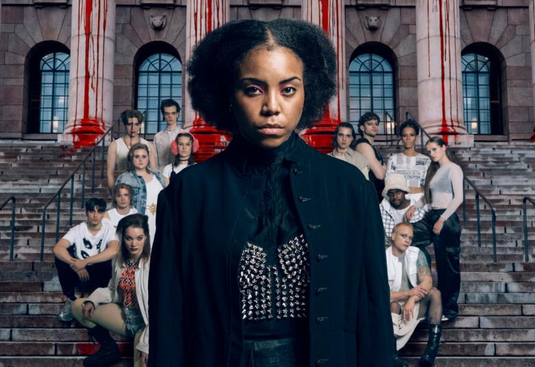 Kuvassa on keskellä nuori nainen mustassa kaavussa, jossa on hopeinen perhonen edessä. Hänellä on mustat, kiharat hiukset. Taustalla on valoisissa puseroissa istumassa harmailla portailla joukko nuoria. 