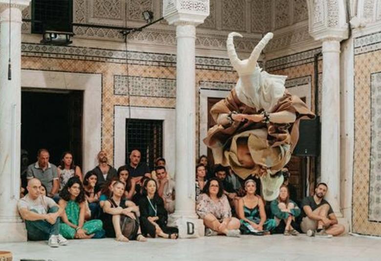 Kuvassa on joukko ihmisiä istumassa rakennuksen valkoisella lattialla ryhmänä ja edessä on kolme valkoista, itämaista pylvästä. taustalla näkyy seinää, jossa on valkoinen, avoin oviaukko ja vieressä kaksi pienempää aukkoa. Seinässä on keltakuvioista mosaiikkia ja sinikuvioista ornamenttia. 