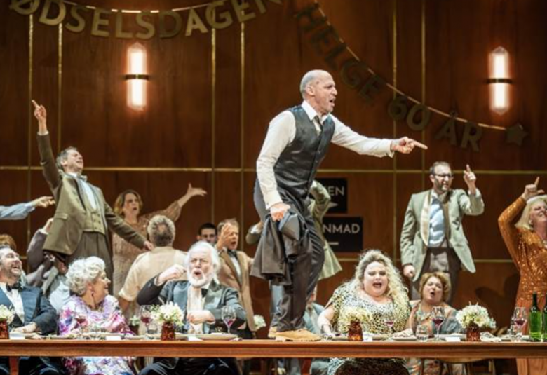 Kuvassa on ihmisiä puvuissa juhlimassa pitkän pöydän ääressä ja takana on juhlivia häenkilöitä kädet levitettynä. Keskellä on mies seisomassa pöydällä sivuttain ja hänellä on harmaa liivi ja housut sekä valkoinen paita. Taustalla on ruskea seinä.