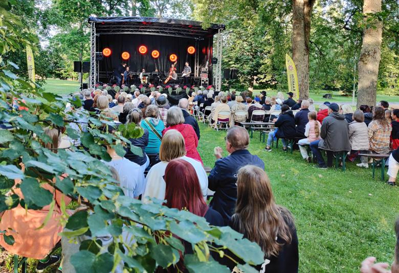 Kuvassa on puiston vihreitä puita ja oksia. Taustalla on esiintymislava ja siinä orkesteri soittamassa. Lavan edestä taakse päin on yleisöä istumassa tuoleilla.