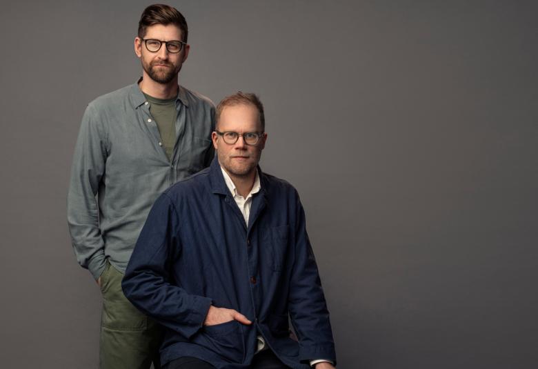 Kuvassa ovat Lars Berge seisomassa harmaassa puserossa Axel Gords Humlesjön takana ja Humlesjö istuu. Hänellä on musta pusero ja pitkät housut. Tausta on tumman harmaa.