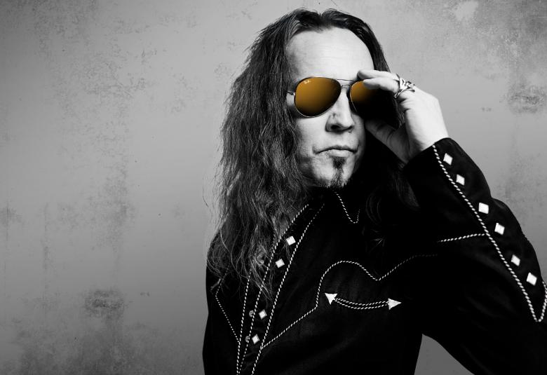 Kuvassa on Jukka ahola puolivartalokuvassa ja hänellä on musta takki päällään. Siinä on nappeja hihassa ja Ahola pitää vasemmalla kädellään aurinkolaseista kiinni. Kuva on musta-valkoinen tausta on harmaa.