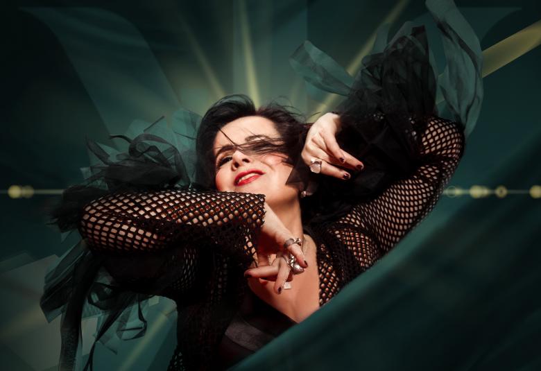 Kuvassa on Kaija Koo puolivartalokuvassa ja hänellä on kädet kohotettuina vauhdikkaasti. Hänellä on musta puku ja hihat ovat verkkomaiset. Tausta hihreän tumma ja valoa tulee pään yläpuolelta.