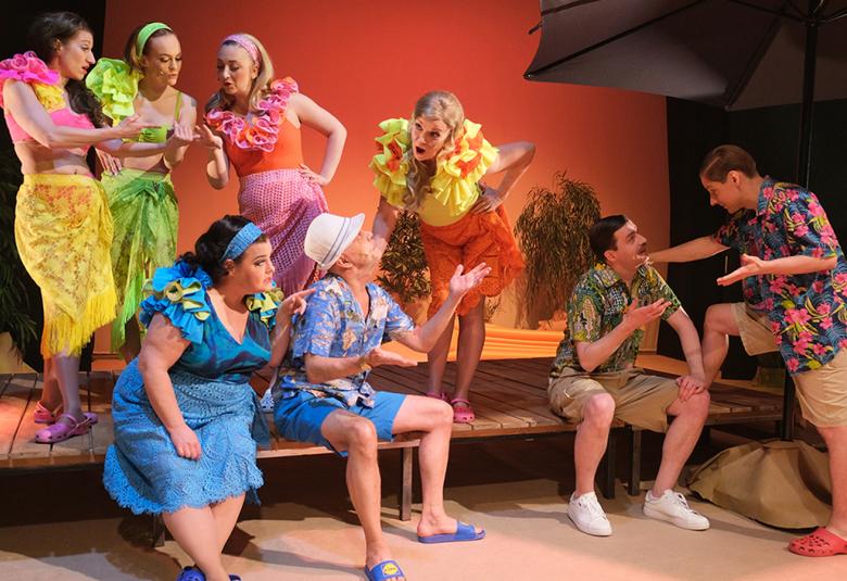 Kuvassa on hieman ylempänä olevalla terassilla seisoo neljä naista ja heillä pastillin värisiä rantamekkoja ja röyhelöt kaulassa. Alempana istuvat sinisessä mekossa nainen ja sinisessä karttapaidassa sekä sinisissä shortseissa mies. Oikealla on kaksi miestä juttelemassa ja heillä on vihertävän sävyiset shortsit ja paidat.