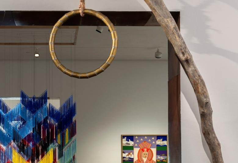 Kuvassa on etualalla kapeaa puunrunkoa ja siitä roikkuu puinen, pyöreä hirttosilmukka. Taustalla näkyy roikkuva tekstiilityö, jossa on Lapin värejä ja näyttelysalia.