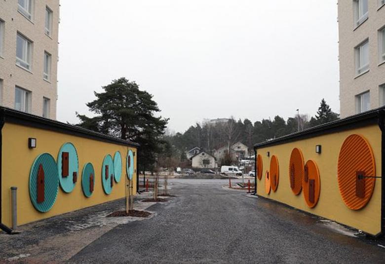 Kuvassa on molemmissa reunoissa vaalean ruskeat kerrostalot. Vasemmalla on pitkä alarakennus ja siinä ruskehtavalla pohjalla on isoja pyöreitä turkooseja alustoja, joilla on tummia yksityiskohtia. Toisella puolella sisääntulotie ja sen toisella puolella vastaava alarakennus ja siinä punaiset pyöreät koristeet. 