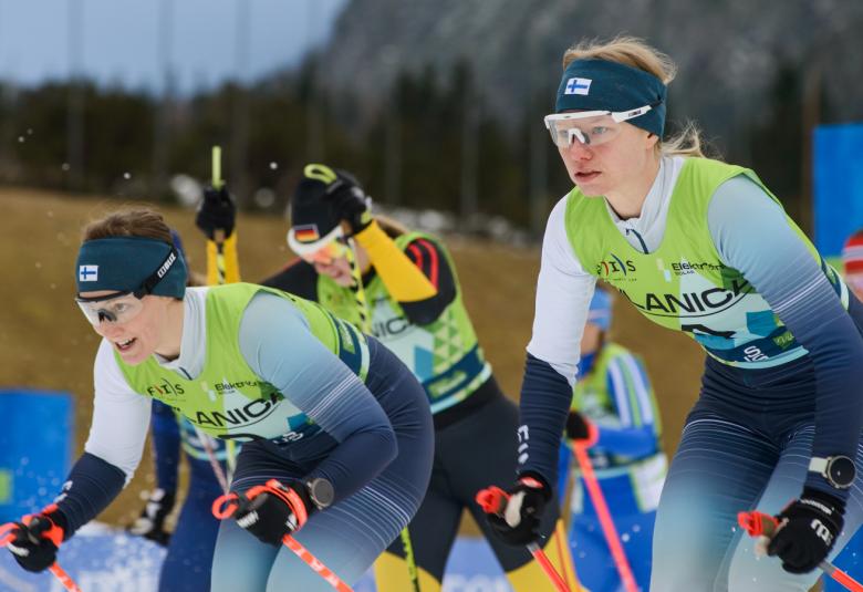 Kuvassa on kolme naishiihtäjää lähdössä kilpailemaan. Heillä on Suomen sini-valkoiset edustusasut ja pipot, joissa on Suomen lippu. 