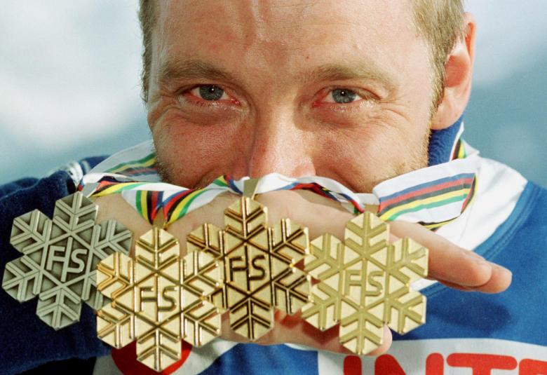 Kuvassa on taustalla Mika Myllylän kasvot ja hän pitää puoliksi kasvojen edessä neljä mitalia, joissa on havukuviota ja keskellä kirjaimet FS. taustalla vaalean sinistä pilveä.