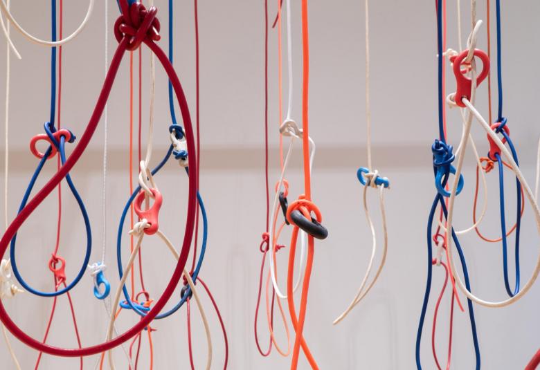 Kuvassa on roikkumassa erivärisiä naruja ja niissä on solmuja. Joihinkin on solmittu iso silmukka. Tausta on harmaa.