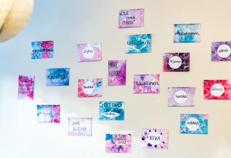 Kuvassa on vaalean sinisellä ja vaalean keltaisella pohjalla lappuja seinällä ja niiden värit ovat puna-sini-valkoisia.  Niissä on tekstejä kuten kiva, rohkea, herkkä jne.