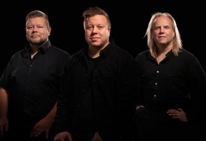 Kuvassa ovat seisomassa kolme miestä ja heillä on mustat puserot päällään. Tausta on myös musta ja kuvassa näkyvät selkeästi kasvot. 