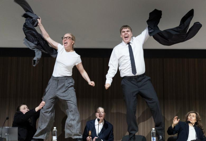 Kuvassa on nuori nainen vasemmalla ja hän heiluttaa takkiaan ja hänellä on valkoinen t-paita ja harmaat, löysät housut päällää, Oikealla on nuori mies, jolla on valkoinen paita jamusta kravatti sekä mustat housut päällään. Taustalla alhaalla on kolme henkilöä pöydän ääressä mustissa takeissa.