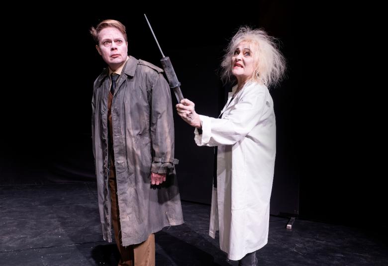 Kuvassa on mies ruskeassa päällystakissa ja oikealla nainen vakoisessa päällystakissa. Tausta on tumma.