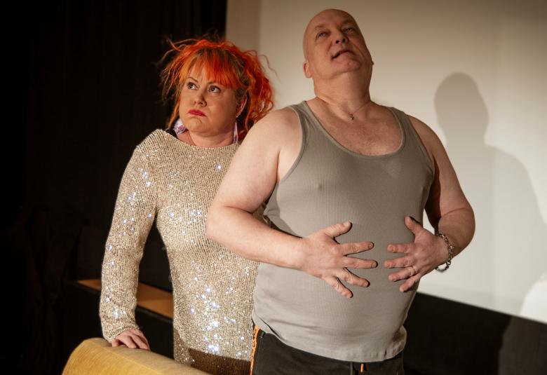 Kuvassa on vasemmalla vaaleassa glitterpuserossa Mira Lukkari seisomassa ja nojaamassa sohvan selkämykseen oikealla kädellään.  Hänen edessään on Jari Lukkari seisomassa harmaassa hihattomassa t-paidassa ja taputtaa oikealla kädellään vatsaansa. 