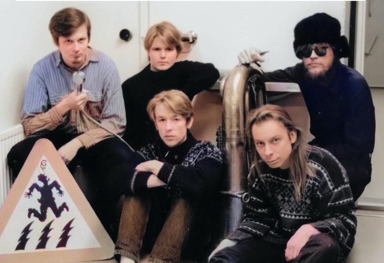 Kuvassa on viisi nuorta miestä, joista kolme istuu takarivissä ja heistä yhdellä on vaalean sininen pusero ja muilla mustat puserot. Edessä istuu kaksi miestä ja vasemmalla heidän vieressään on kolmioliikkennemerkki jossa on pomppivan henkilön kuva. Kuva on osa kirjan kannesta.