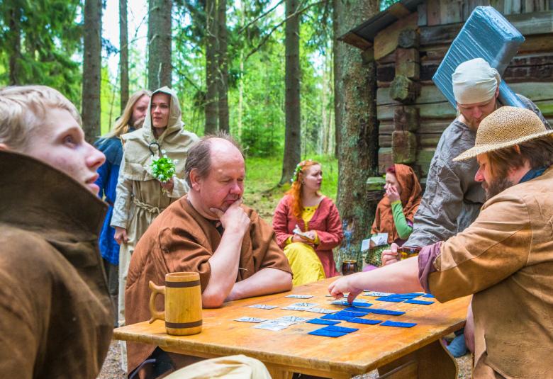 Kuvassa on metsässä olevan harmaan hirsimökin vieressä pöydän ääressä vastakkain kaksi miestä ja pöydällä on sinisiä kortteja, joita katsovat. Pöydän päässä seiroo nainen harmaassa huivissa ja vasemmassa kulmassa ruskeassa takissa nuori mies. Taustalla näkyy ihmisiä keskiaikaisissa vaatteissa.