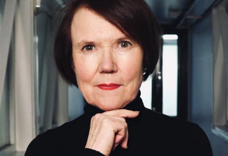 Kuvassa on Rosa Liksom puolivartalokuvassa mustassa poolopuserossa ja hänellä on oikea käsi leuan alla. Hänellä on tummat, punertavat puolipitkät hiukset. Taustalla on harmaata käytävää.