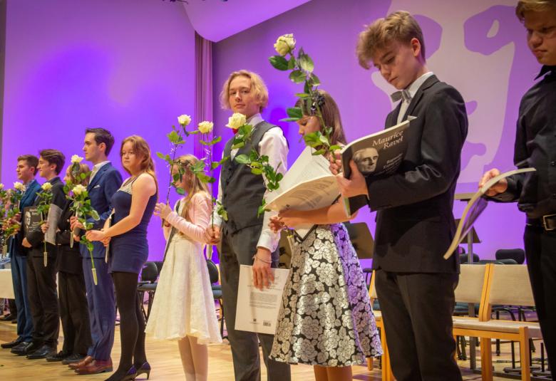 Kuvassa on viistosti seisomassa näyttämöllä sekä tyttöjä että poikia. Heillä on kädessään ruusu ja kunniakirja. Taustalla on violettia valoa.
