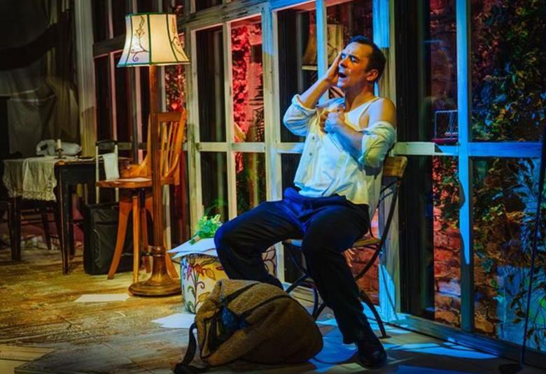 Kuvassa on Ilja Peltonen kalustetussa huoneessa, jossa Ilja Peltonen istuu tuolilla ja nojaa taaksepäin.  Hänellä on vaalean sininen paita ja siniset housut päällään. Huoneessa on pieni pöytä ja iso jalkalamppu sekä takana isot ikkunat. 