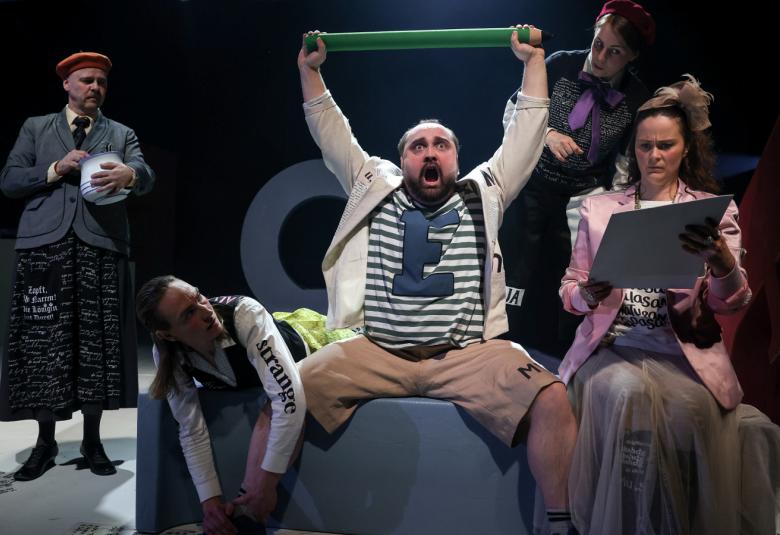 Kuvassa on keskellä Sami Sainio, joka istuu sohvalla ja pitää vihreää keppiä ylhäällä käsissään.  Vieressä istuu nainen vaaleassa takissa. Hänellä on kansia kädessään. Taustalla seisoo harmaassa takissa nainen.