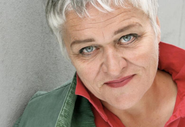 Kuvassa on Pirkko Saision kasvokuva ja hänellä näkyy kuvassa harmaata takkia ja punaisia kauluksia. Hänellä on vaalean harmaat hiukset.