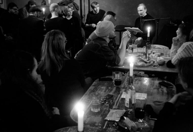 Kuvassa on huone täynnä ihmisiä. Oikealla on pitkä pöytä ja sen äääressä on ihmisiä juomassa ja keskustelemassa. Pöydässä on kynttilöitä. Vasemmalla on ihmisiä seisomassa. Kuva on musta-valkoinen.