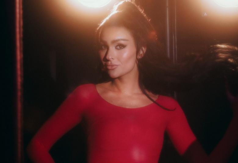 Kuvassa on keskellä Sara Siipola puolivartalokuvassa. Hänellä on punainen mekko päällään ja hänellä on tummat hiukset nutturalla. Taustalla tumman ruskeaa ja kaksi ylävaloa.