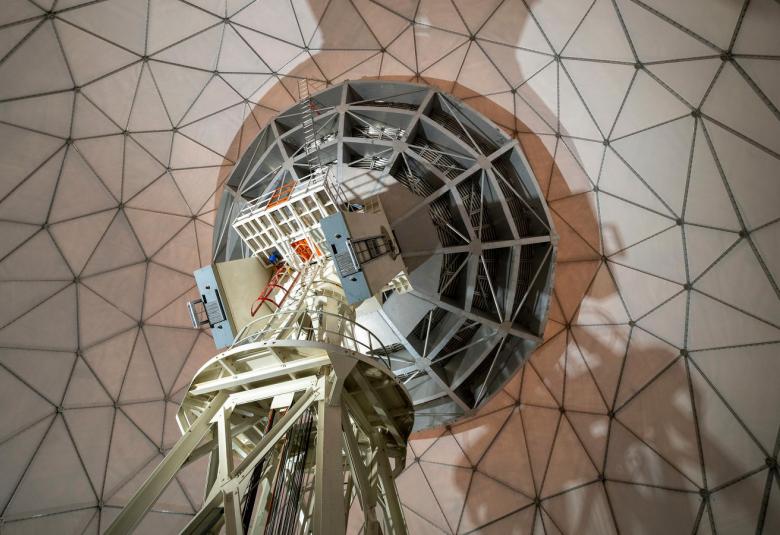 Kuvassa on avaruusteleskooppi kuvattuna alhaalta ylöspäin ja taustallaon rautaverkkoa vaaleanruskealla pohjalla.