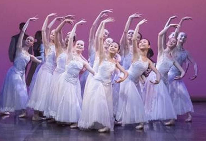 Kuvassa on iso joukko nuoria ballerinoja valkoisissa, pitkissä mekoissa oikea käsi ylhäällä kaarella. Tausta on vaalea lila.