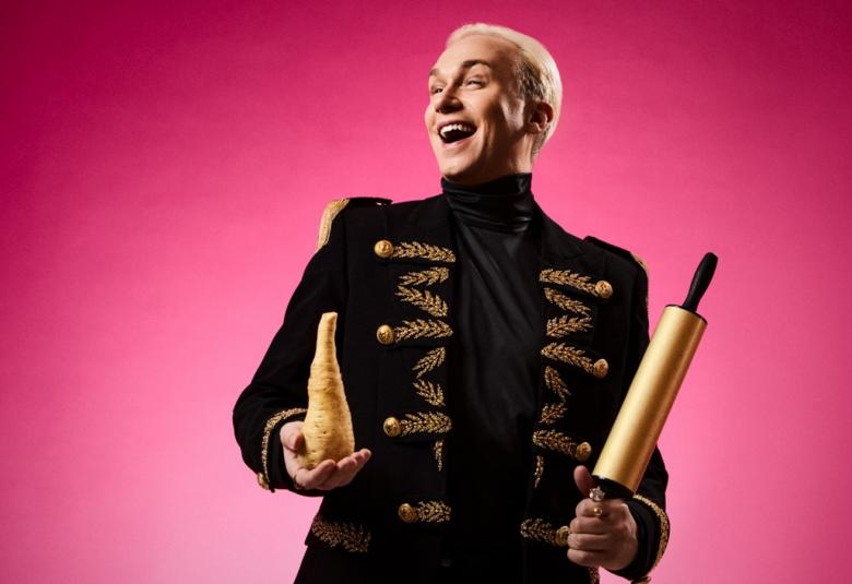 Kuvassa on Christoffer Strandberg mustassa kultakoristellussa takissa ja palsternakka oikeassa kädessään. Takana on pinkkiä taustaa.