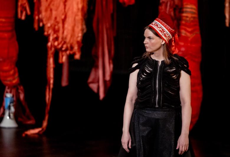 Kuvassa on roikkuvia tumman punaisia tilataideteoksia tummalla taustalla. Oikealla edessä on Elle Sofe Sara seisomassa ja hänellä on musta hihaton mekko ja punainen kirjailtu Lapin päähine päässään. 