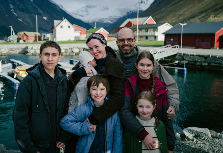 Kuvassa on keskellä  perhe ulkona vuonomaisemassa ja taustalla näkyy tuntureita ja niiden edessä kaksi taloa. Edessä on neljä lasta ja takana isä sekä äiti vauva sylissään.