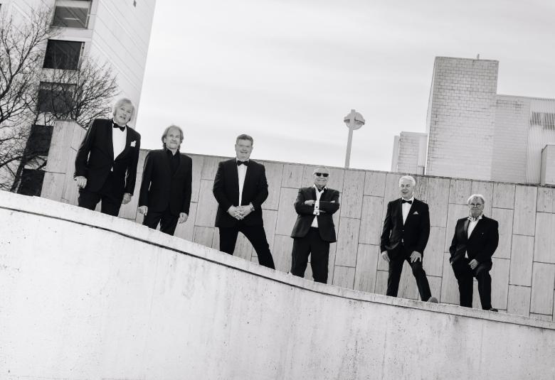 Kuvassa on alaspäin viettävän kaiteen vierellä kuusi mieslaulajaa seisomassa mustissa puvuissa.  Taustalla on vaaleaa seinämää ja vasemmalla kerrostalon julkisivua ikkunoineen. Kuva on musta-valkoinen.