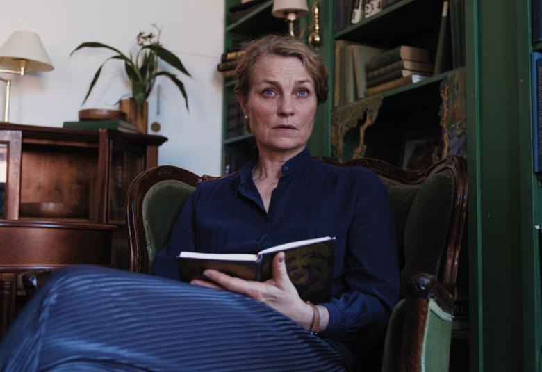 Kuvassa on Jonna Järnefelt istumassa nojatuolissa ja hänellä on kädessään muistikirja kädessään. Takana on ruskea matala kirjahylly ja oikealla on iso tumman ruskea kirjahylly.