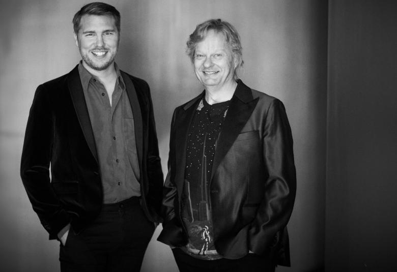 Kuvassa on seisomassa vasemmalla Aarne Pelkonen ja häenllä on musta takki päällään. Oikealla on Santtu-Matias Rouvali myös mustassa takissa. Kuva on musta-valkoinen.