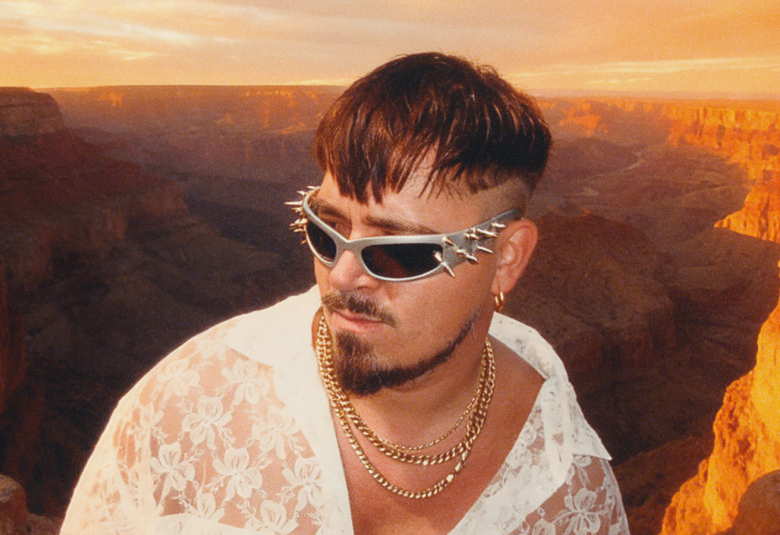 Kuvassa on Käärijä keskellä alempana ja hänellä on valkoinen paita päällään ja aurinkolasit. Taustalla on ilta-auringon valossa olevaa Grand Canonia.