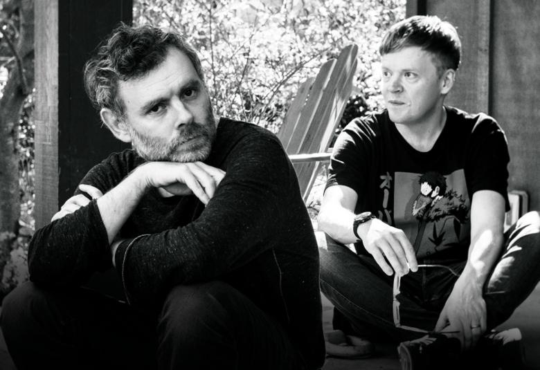 Kuvassa on vasemmalla edessä istumassa kyyryssä Gabriel Kahane ja hänen takanaan oikealla kyykyssä on Pekka Kuusisto. Taustalla seinää ja pensaistoa. Kuva on musta-valkoinen.