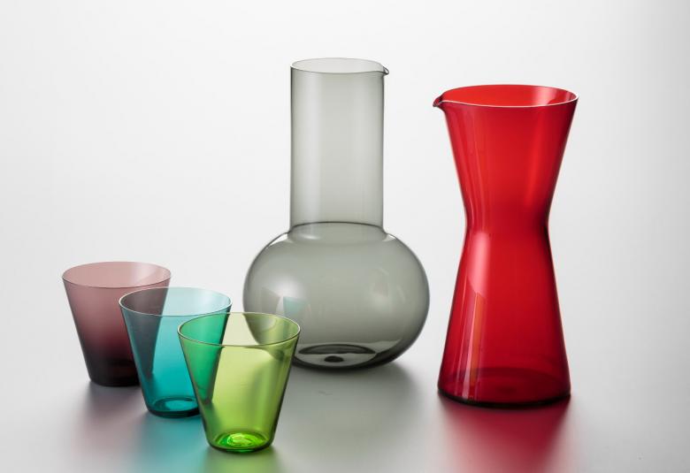 Kuvassa on vasemmalla kolme eri väristä lasia ja keskellä harmaa iso alhaalta pyöristetty maljakko. Oikealla on punainen kaatomaljakko. Tausta on vaalean harmaa.