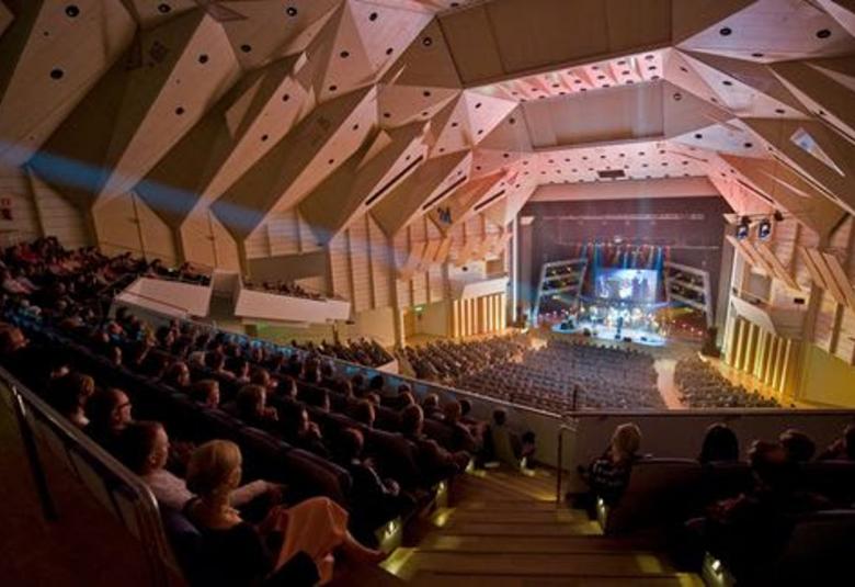 Kuvassa on Tampere-talon konserttisali ylhäältä alaspäin ja hämärässä salissa on yleisöä istumassa  ja katsomassa alhaalla olevalle näyttämölle orkesteria. Vasemmalla ylhäällä on taitettua kattoa.