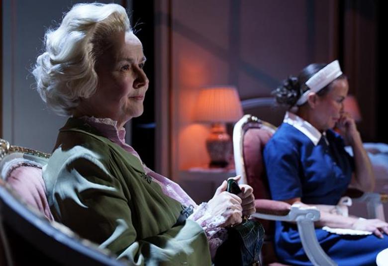 Kuvassa on etualalla Paula Siimes istumassa nojatuolissa vinottain ja hän pitää kävelykepistä kiinni edessään. Taustalla näkyy Maria Kuusiluoma istumassa valkoinen hilkka päässään.