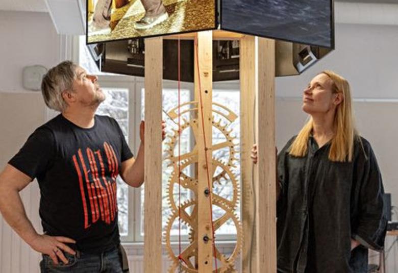 Kuvassa on vasemmalla Oliver Kochta-Kalleinen katsomassa ylöspäin puisen kellokoneiston yläpuolella olevaa neliötä ja sen koneistoa. Oikealla on Tellervo Kalleinen seisomassa ja katsomassa myös ylöspäin. Hänellä on musta leveä mekko. Taustalla on vaalealla seinällä ikkunoita.