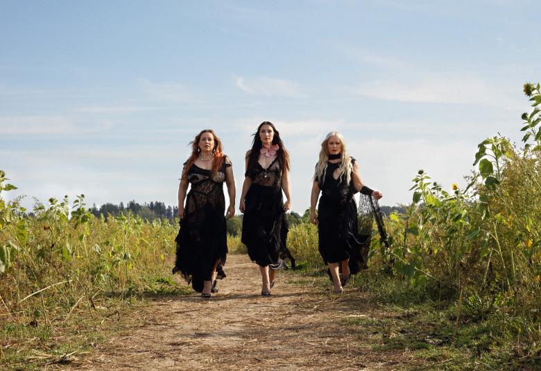 Kuvassa ovat Rodeon kolme jäsentä kävelyllä hiekkatiellä ja vierellä on korkeita kasveja pellolla. Heillä tummat pitkät mekot päällään. Taustalla on sinistä taivasta.